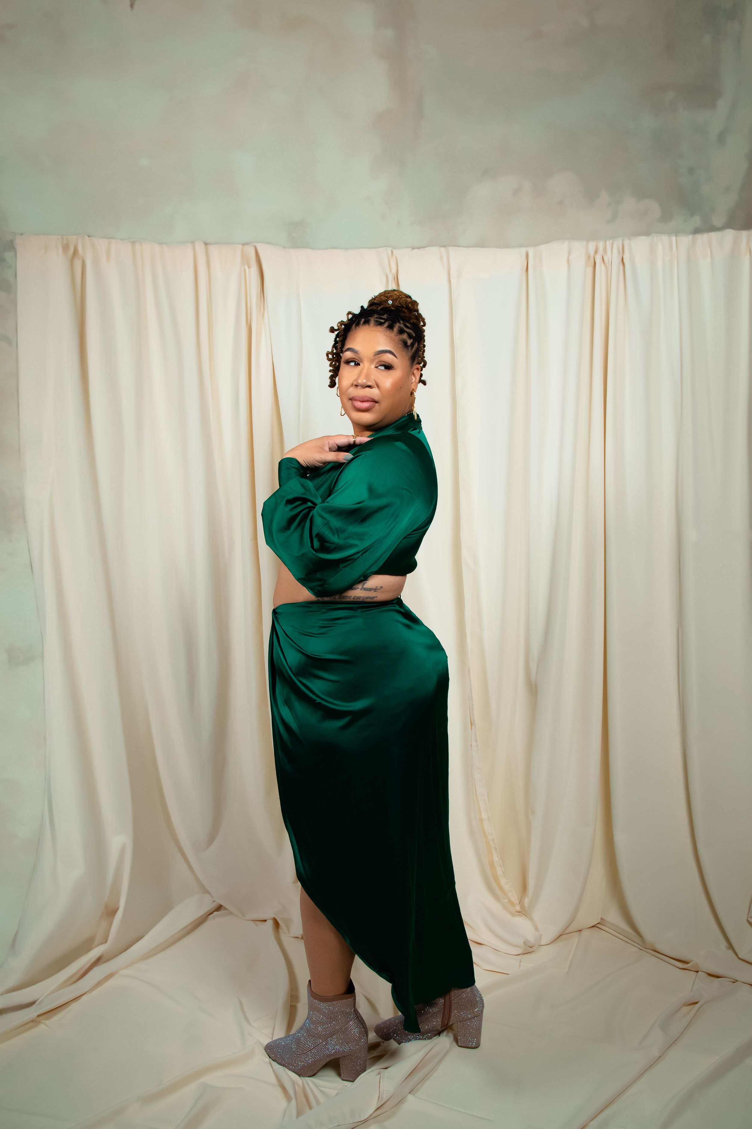 A portrait of someone in an elegant emerald green silk dress against a light backdrop with flowing curtains.