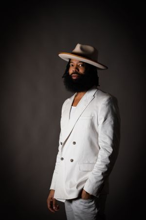 Fashion portrait featuring wide-brimmed hat and white blazer against dark backdrop.