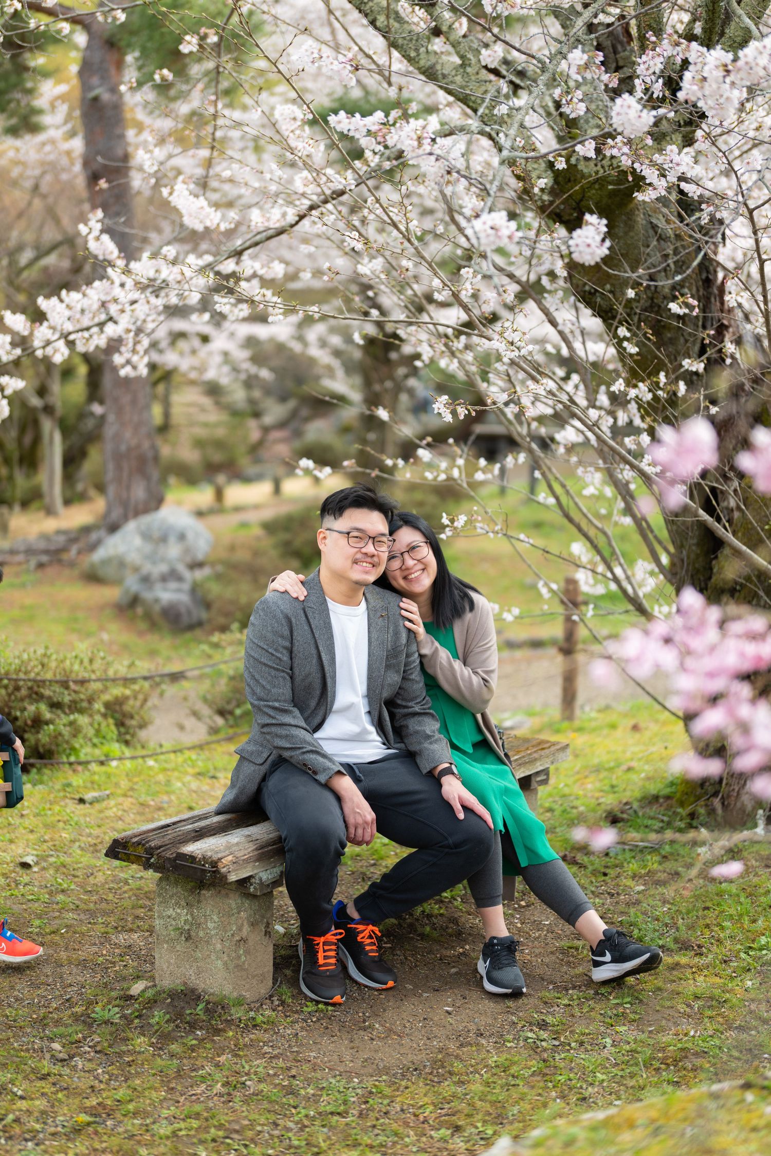 Smiling couple posing on stone bench surrounded by pink and white cherry blossoms.
