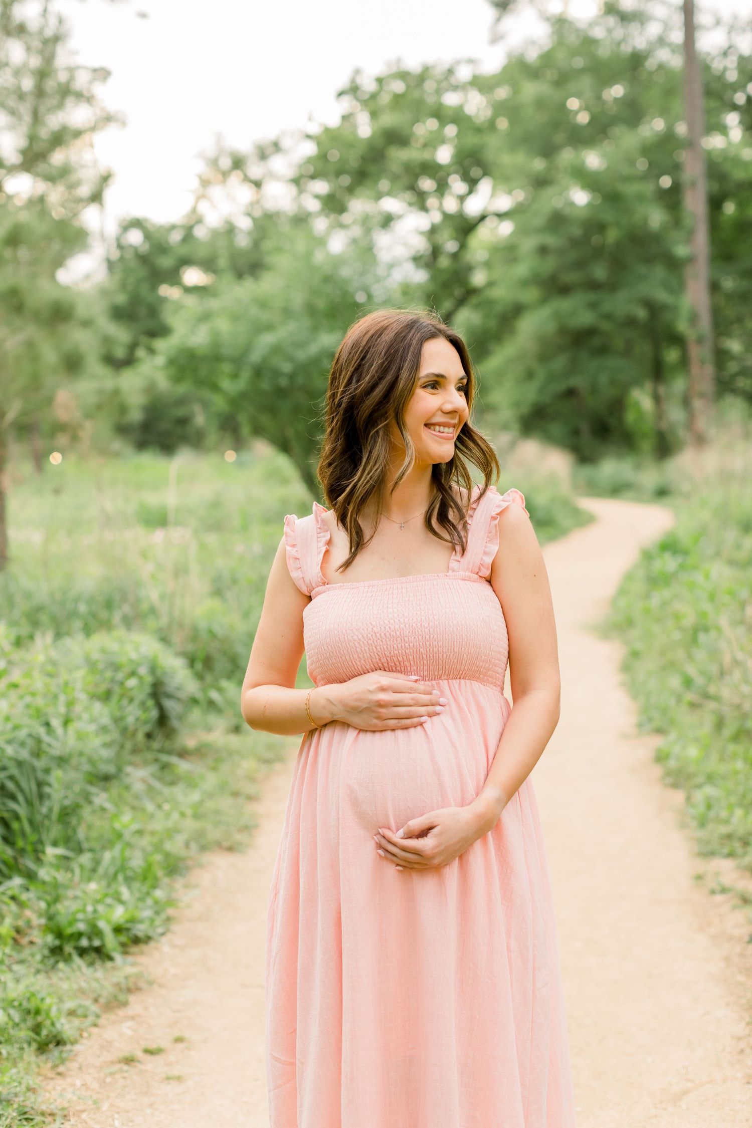A woman in a pink maternity dress stands on a dirt path surrounded by lush greenery.