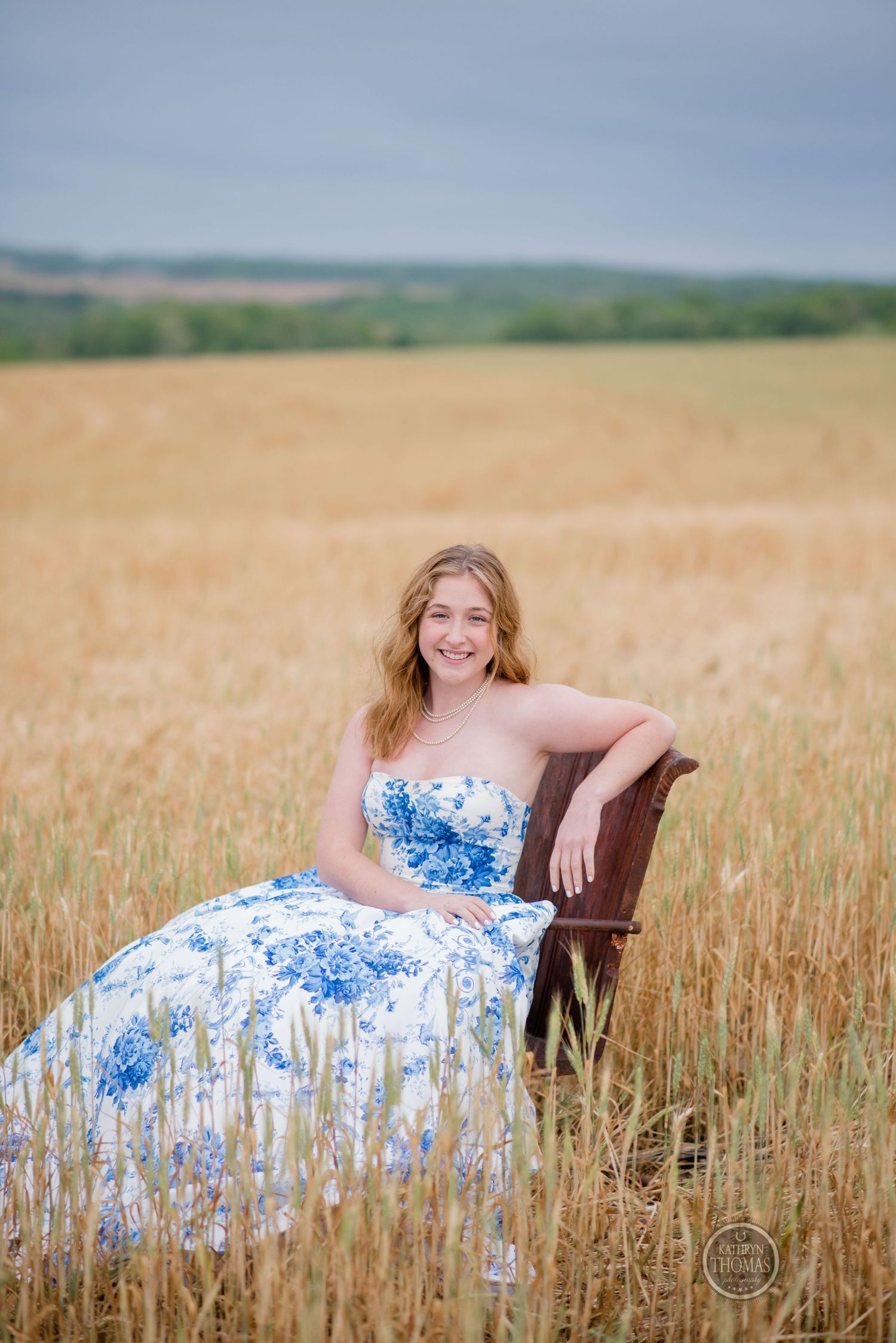 Woman in a blue and white floral gown poses in an antique chair amidst a golden wheat field.