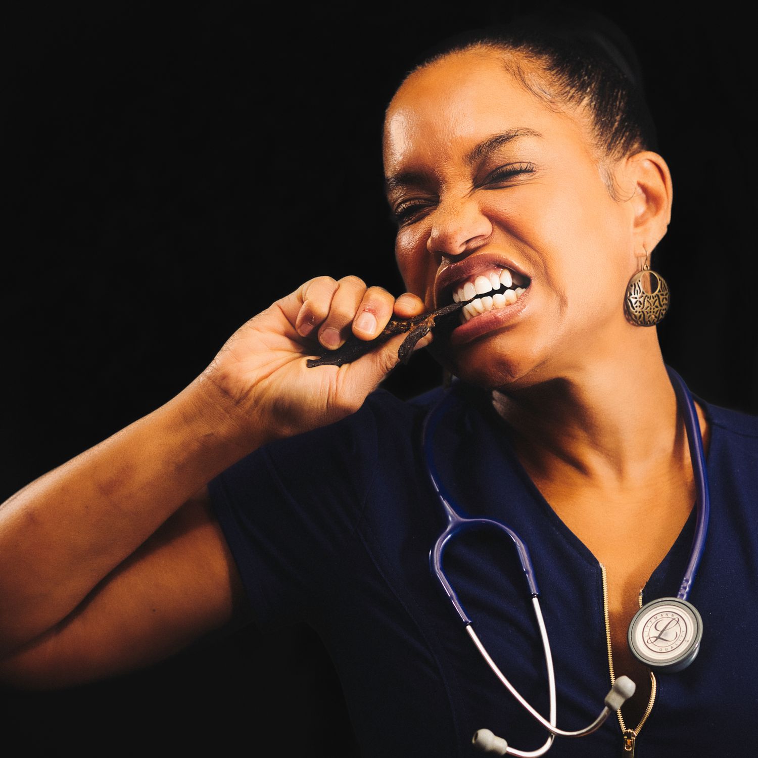 Healthcare professional wearing blue scrubs and stethoscope laughing joyfully against dark background.