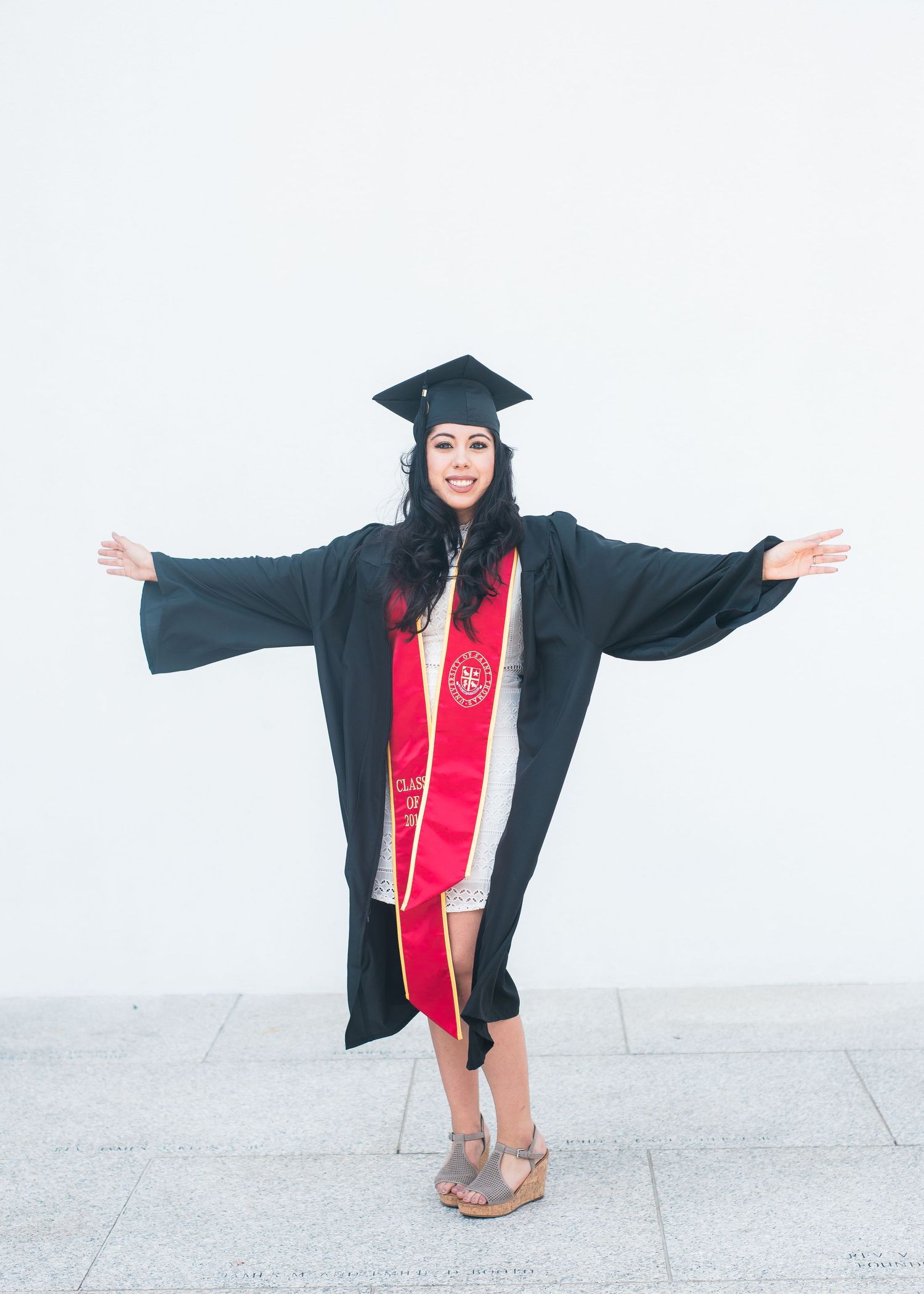 woman raising her arms wearing a black graduation cap and gown and red sash