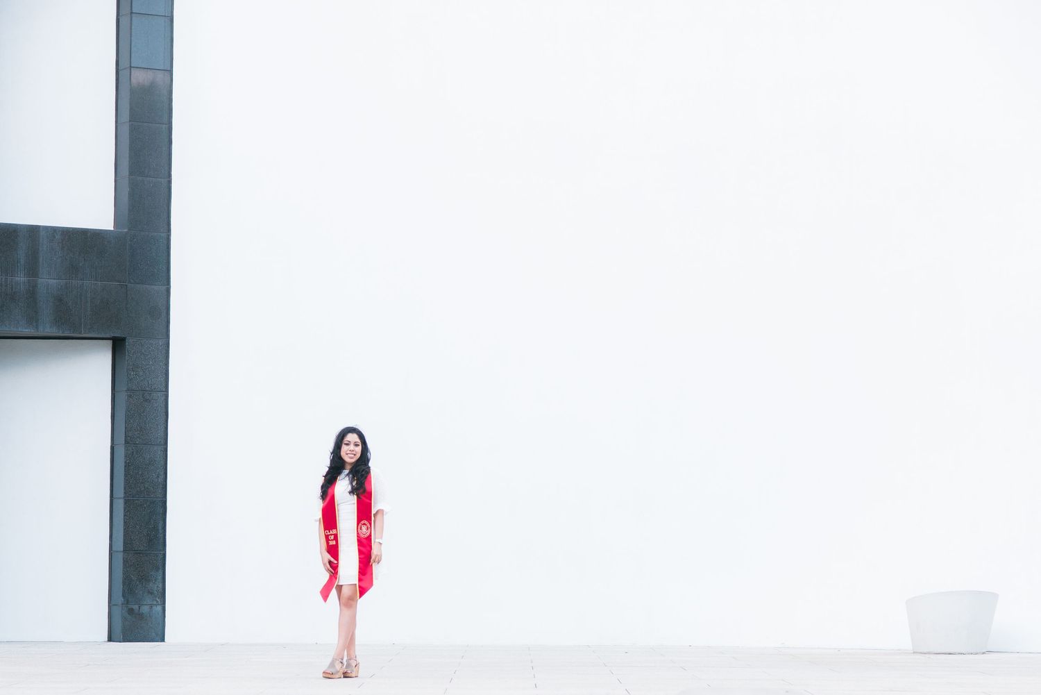 woman standing behind a white wall at University of St. Thomas campus wearing a graduation sash
