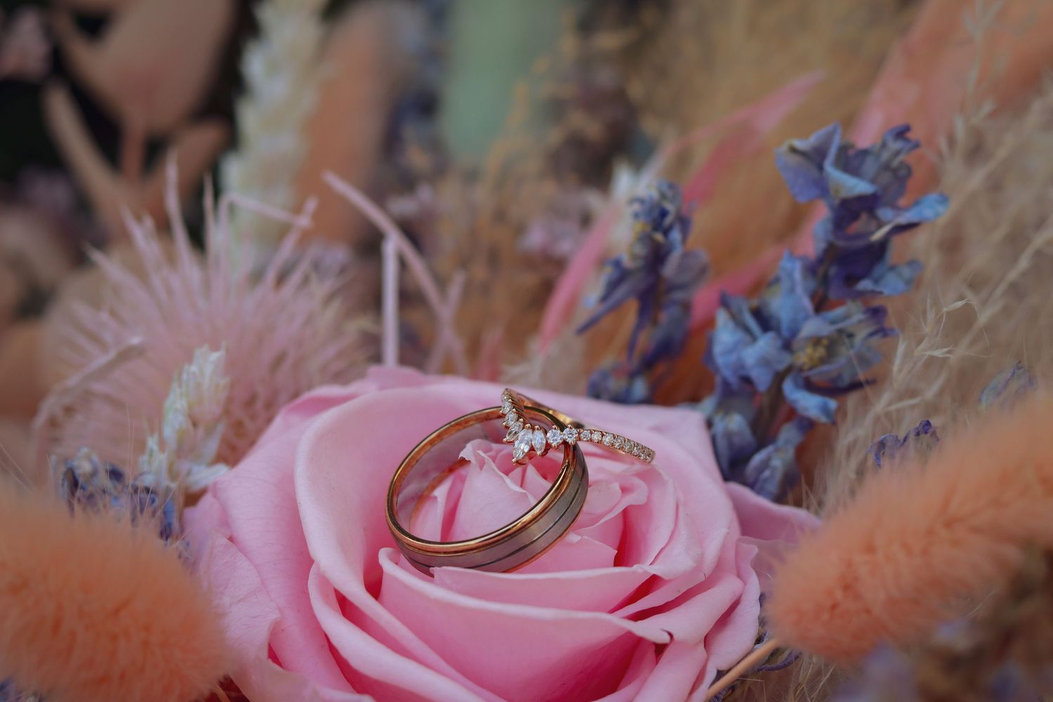 Wedding rings nestled on pink rose with purple flowers and soft pink accents.