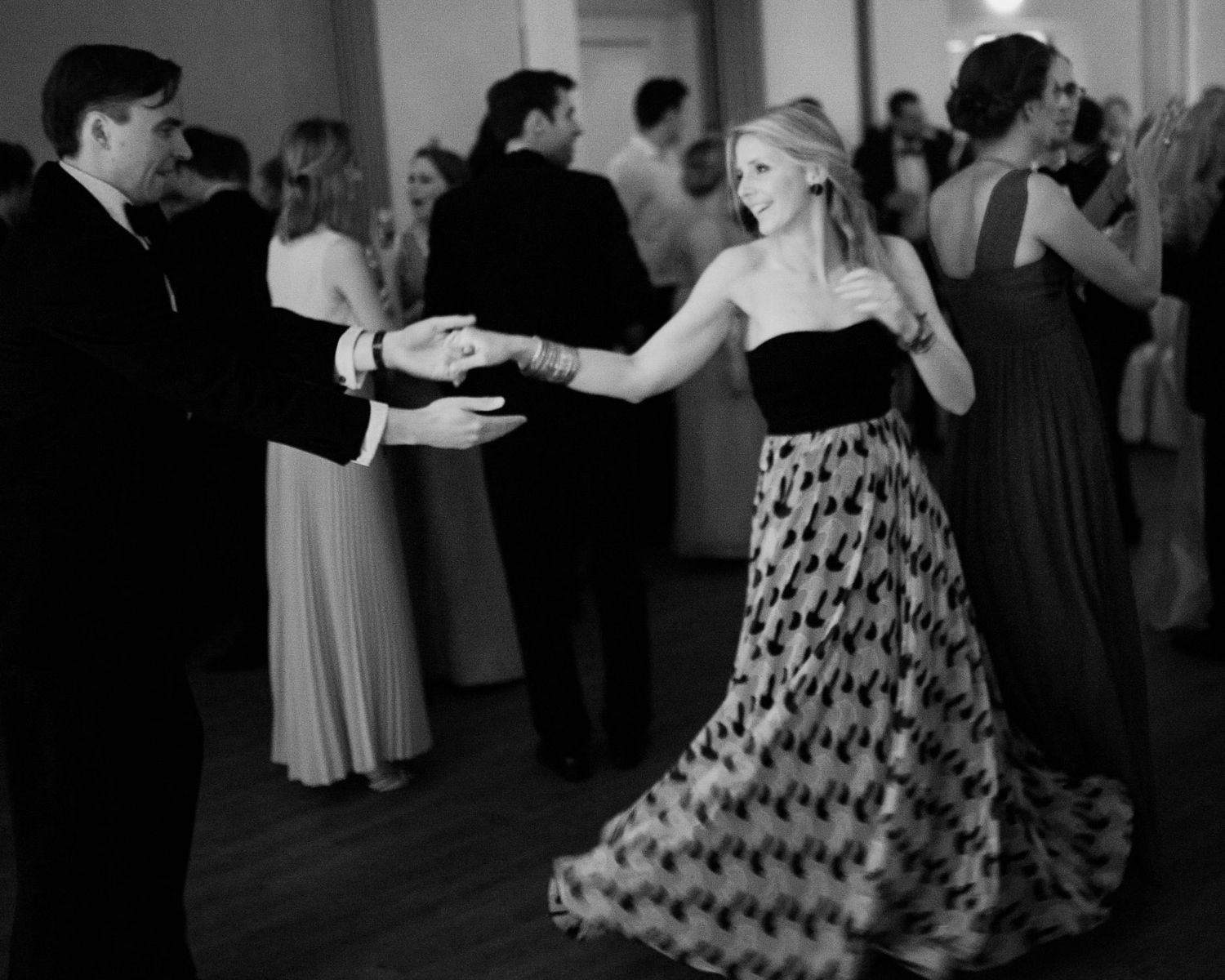 Black and white photo of dancers in formal attire moving gracefully across a ballroom floor.