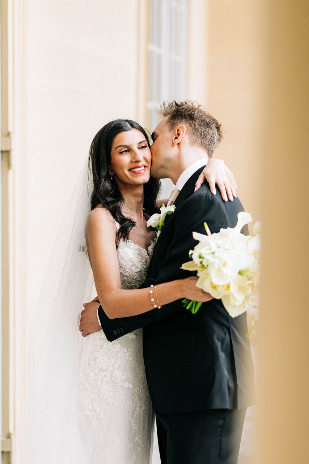 Tender wedding moment captured against a cream-colored wall with white floral bouquet.