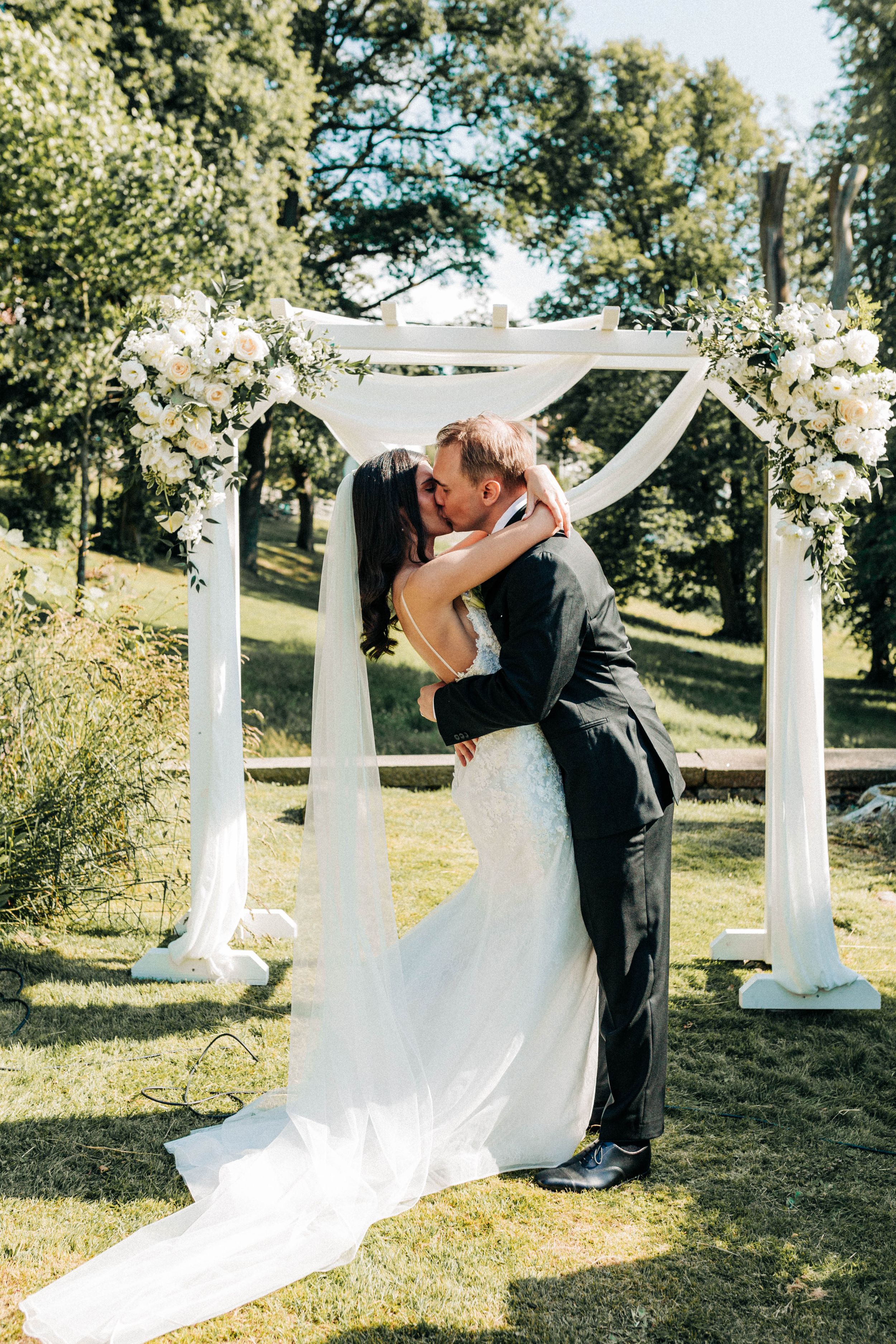 A romantic wedding ceremony under a white floral arch decorated with flowers in a sunlit outdoor garden setting.