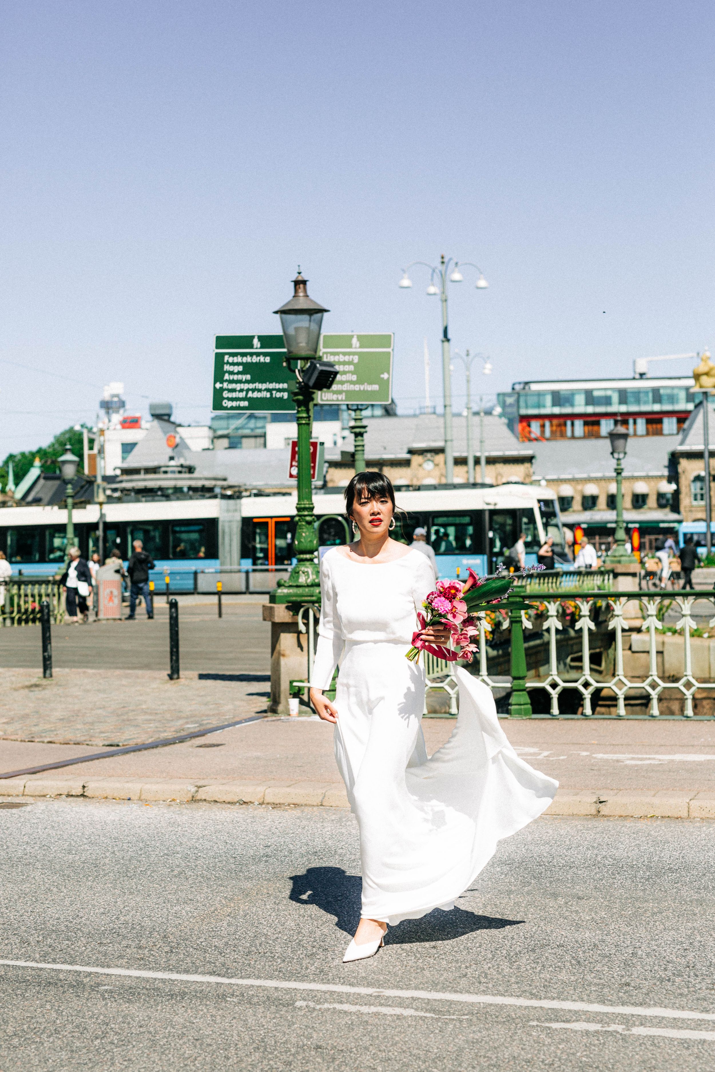 A bride in a flowing white dress walks along a seaside promenade carrying a vibrant bouquet.