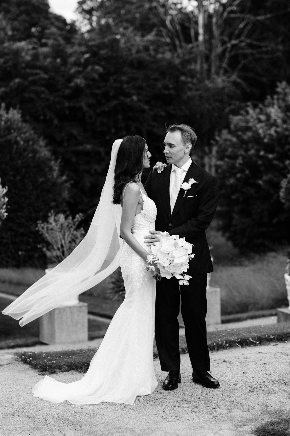Classic black and white portrait of newlyweds outdoors with flowing veil and white bouquet.