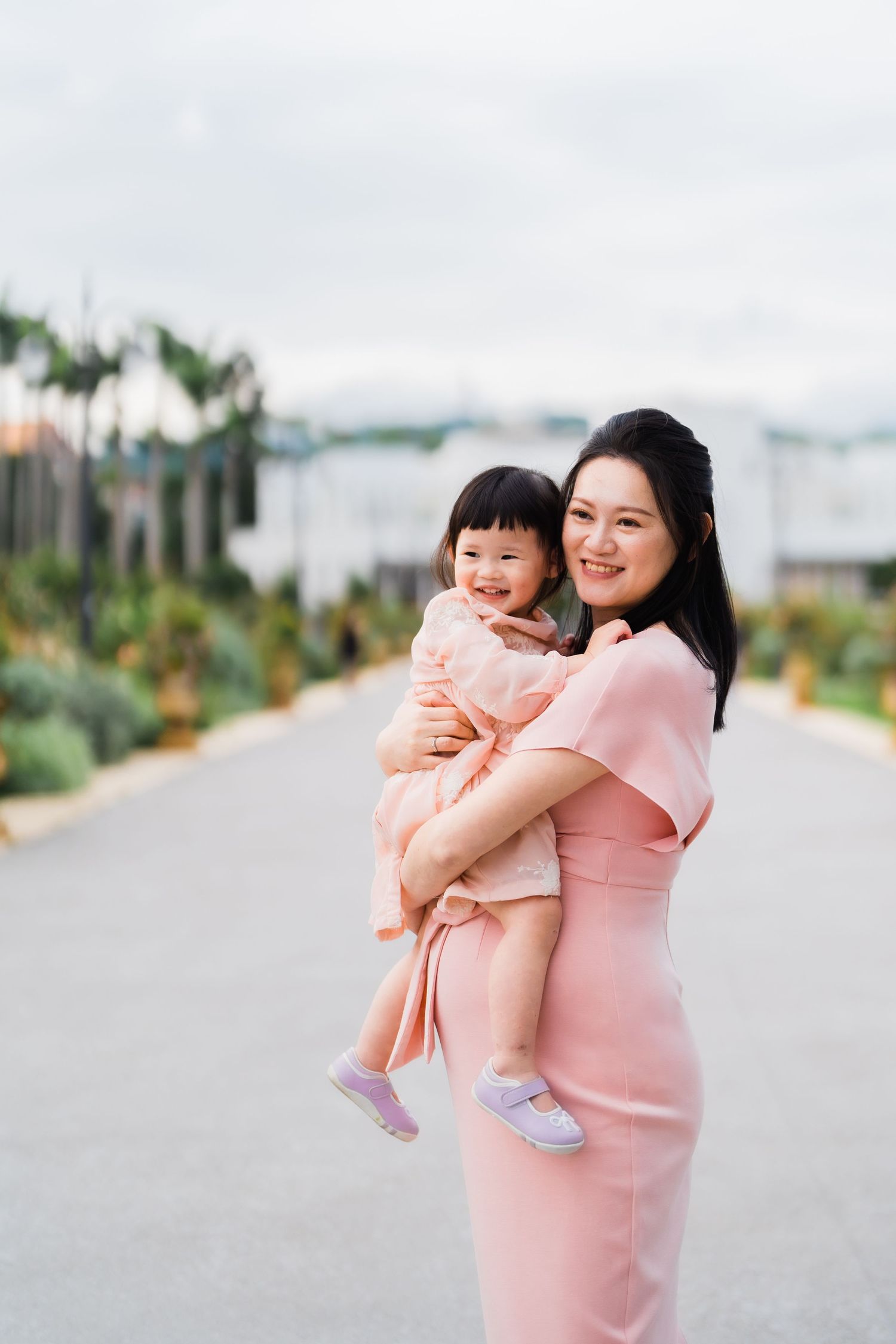 Person in pink dress holding small child on a pathway with palm trees in background.