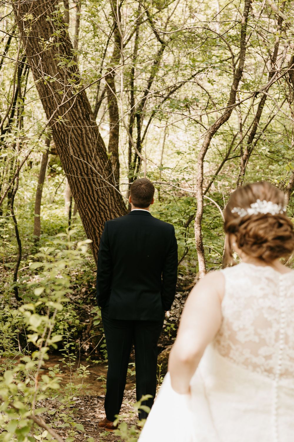 Groom in black suit stands facing forest trees as bride in lace wedding dress approaches from behind.