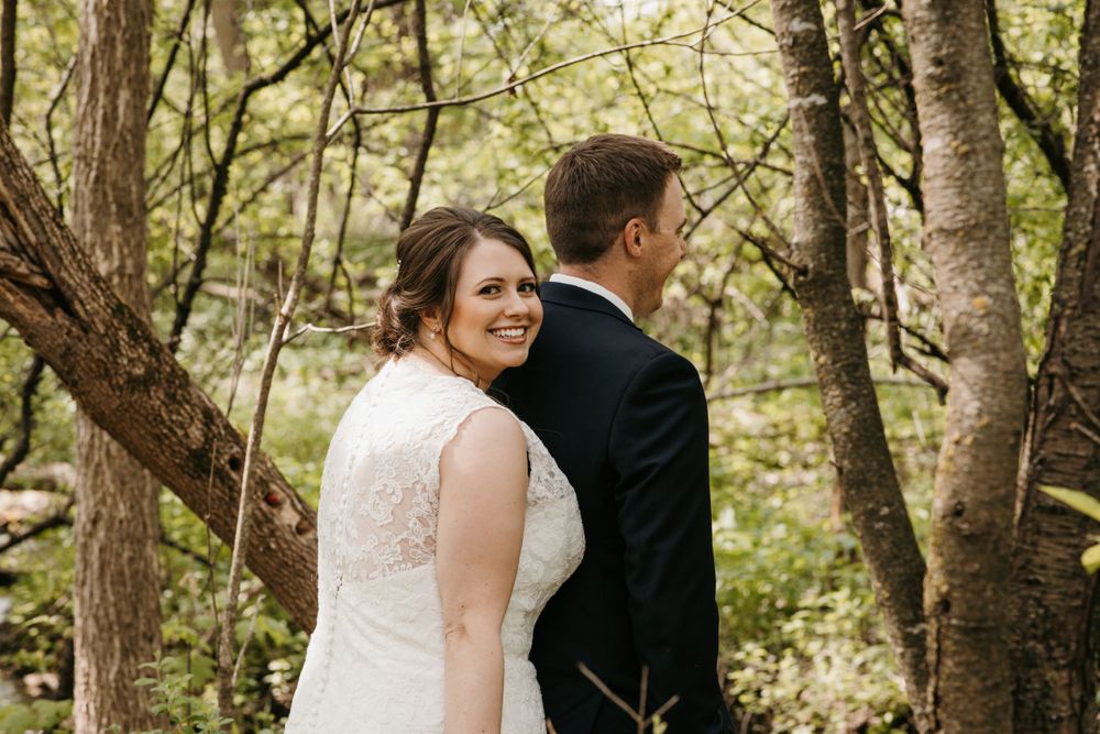 Bride in lace gown smiles back at groom while standing among trees in a forest.
