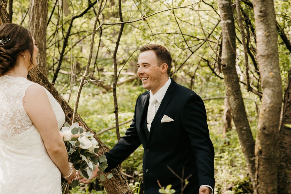 Groom in black suit smiles joyfully at bride holding white bouquet during first look in lush forest.