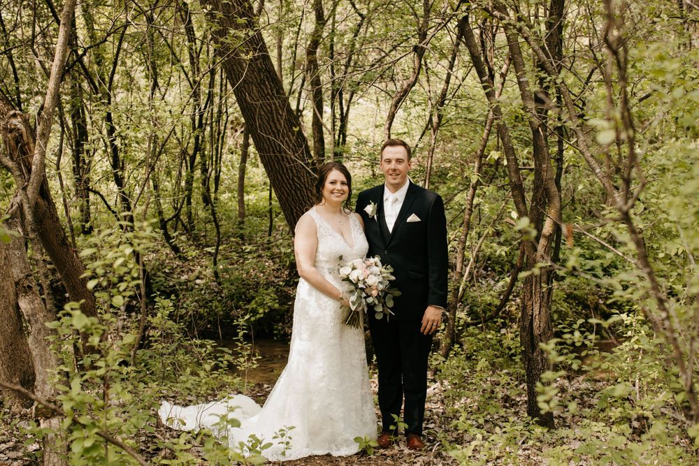 Bride and groom pose together in lace wedding gown and black suit holding bouquet in wooded setting.