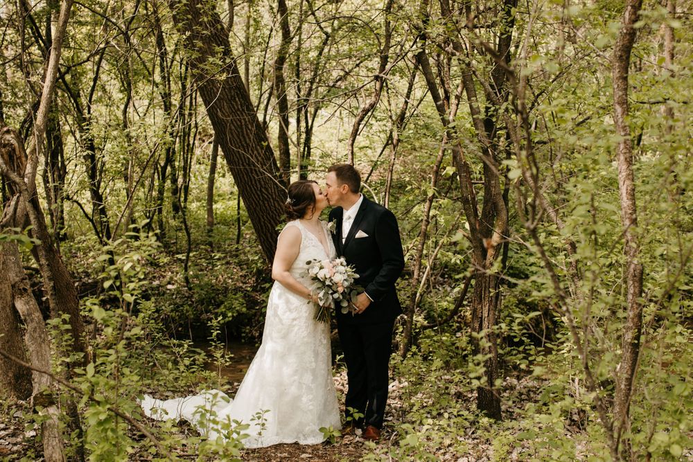 Bride and groom share a romantic kiss surrounded by lush green trees in a woodland setting.