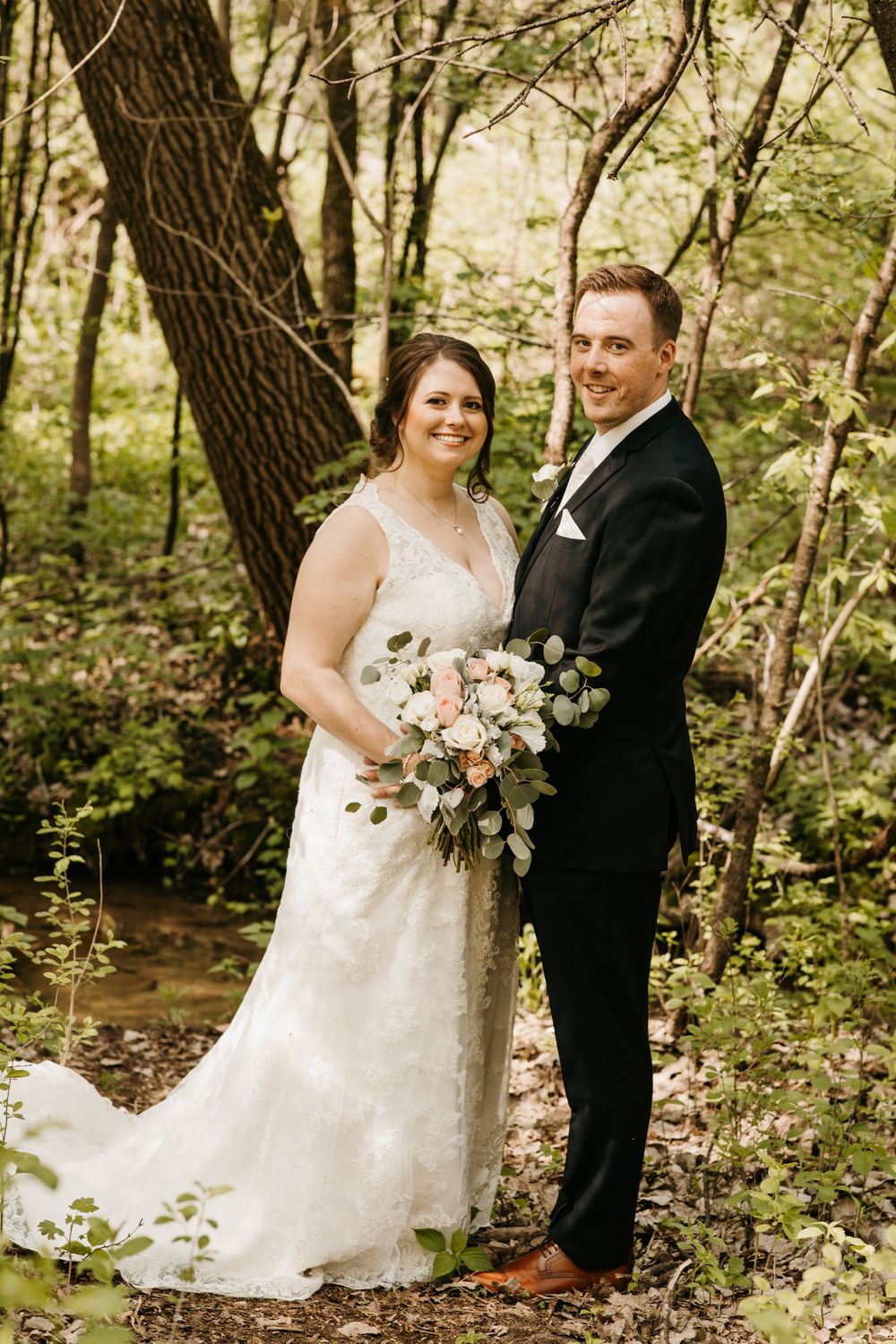Smiling bride and groom pose in forest, bride wearing lace gown and holding pink and white bouquet.