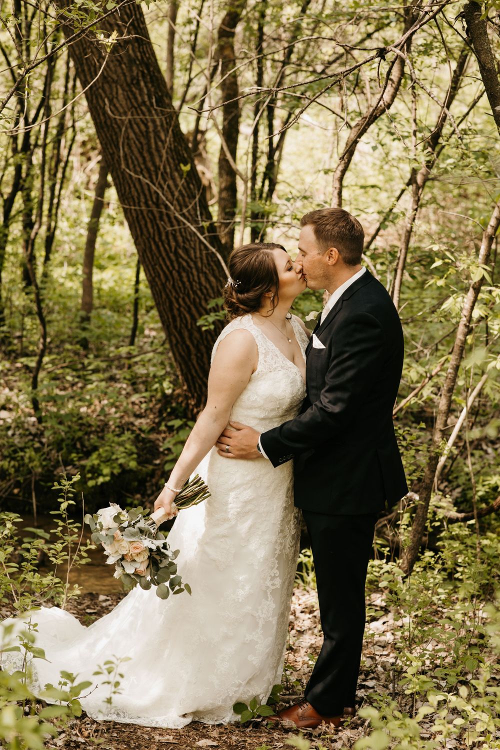 Bride and groom share a tender kiss in a sunlit forest, bride holding a white and pink bouquet.