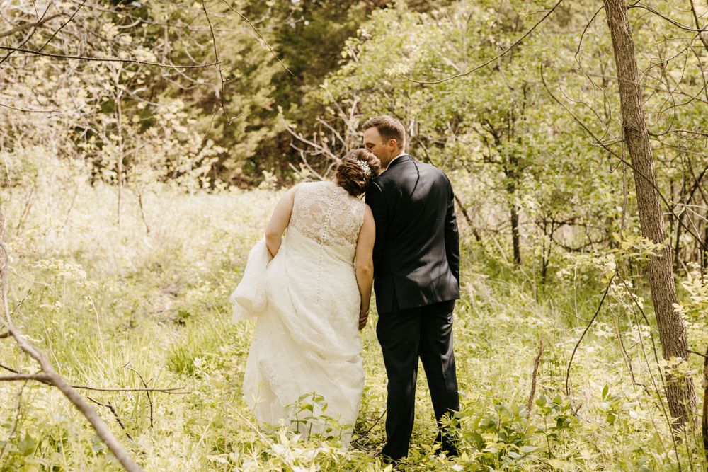 Bride in lace gown and groom in black suit walk hand in hand through a bright green forest.