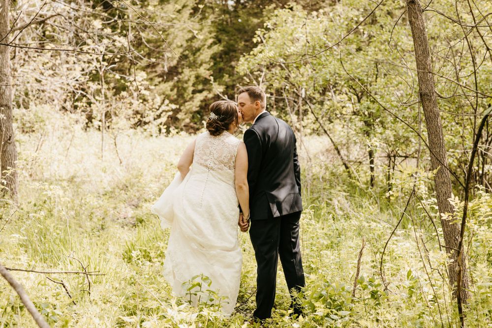 Bride and groom share a romantic kiss while holding hands in a sunlit green woodland setting.