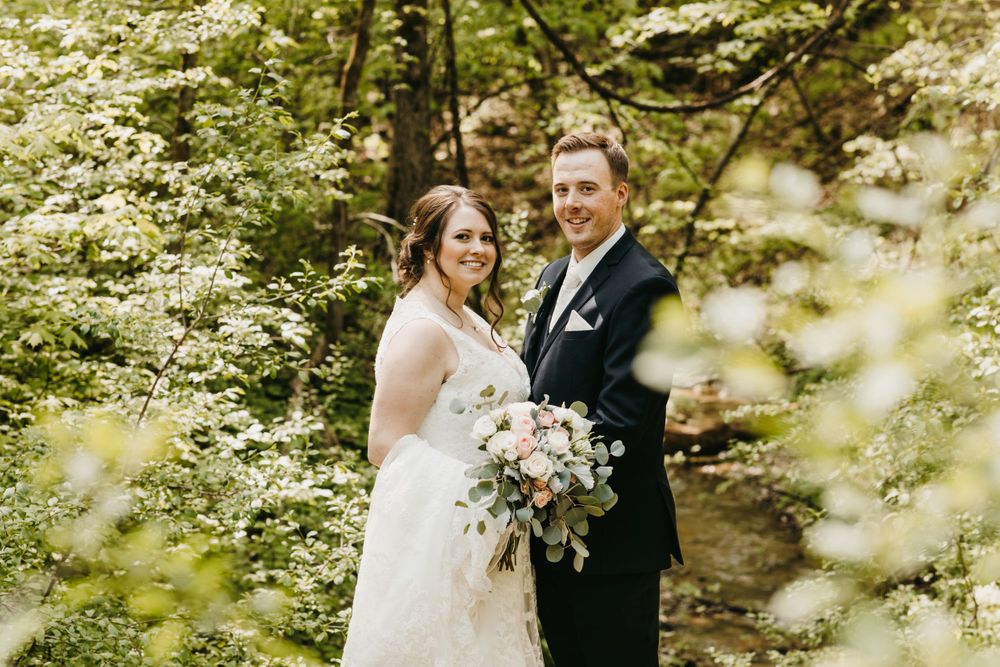 Smiling bride and groom pose among blooming white trees, bride holding an elegant floral bouquet.