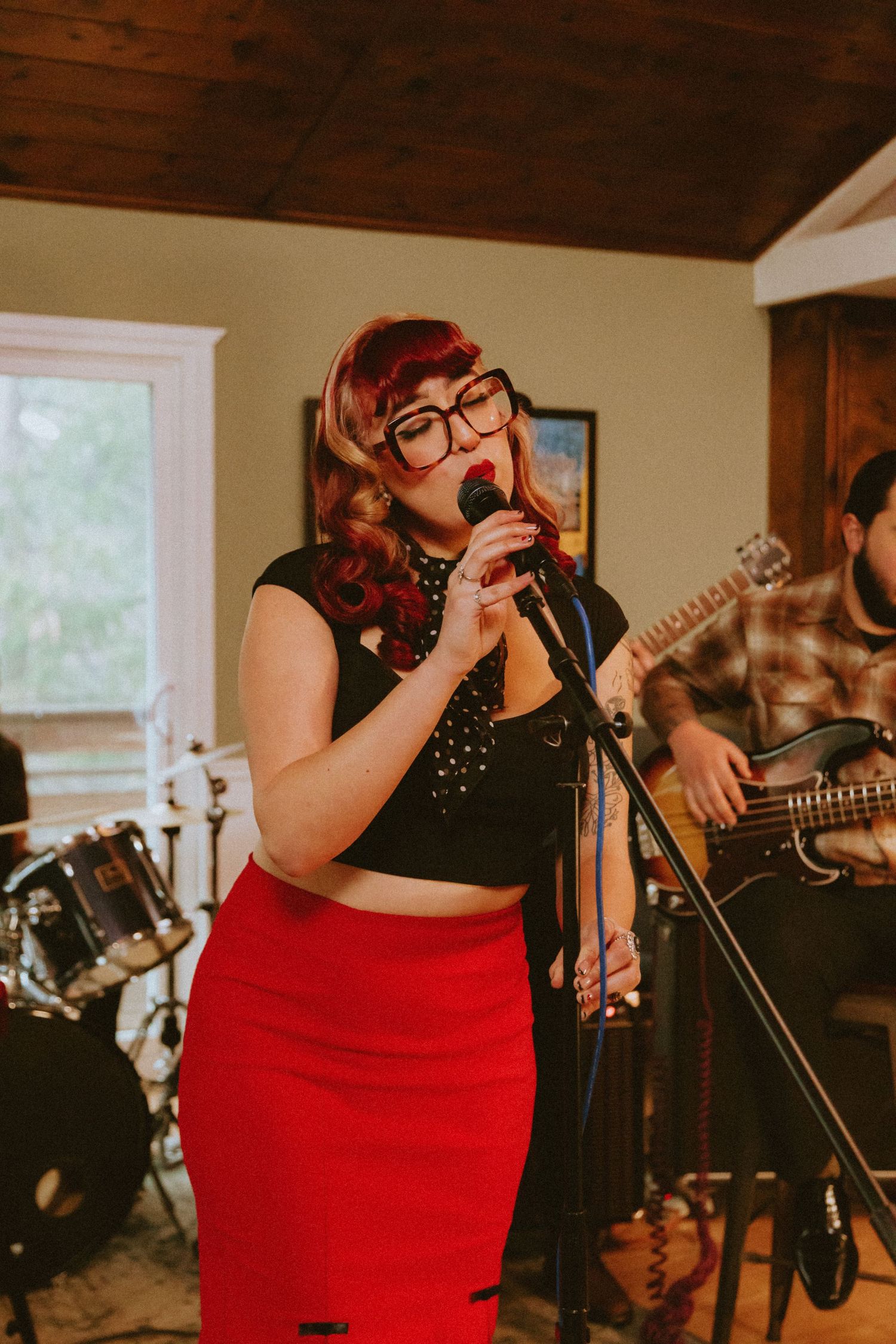 Live band performing in a cozy home setting with vocalist in red skirt and black top.
