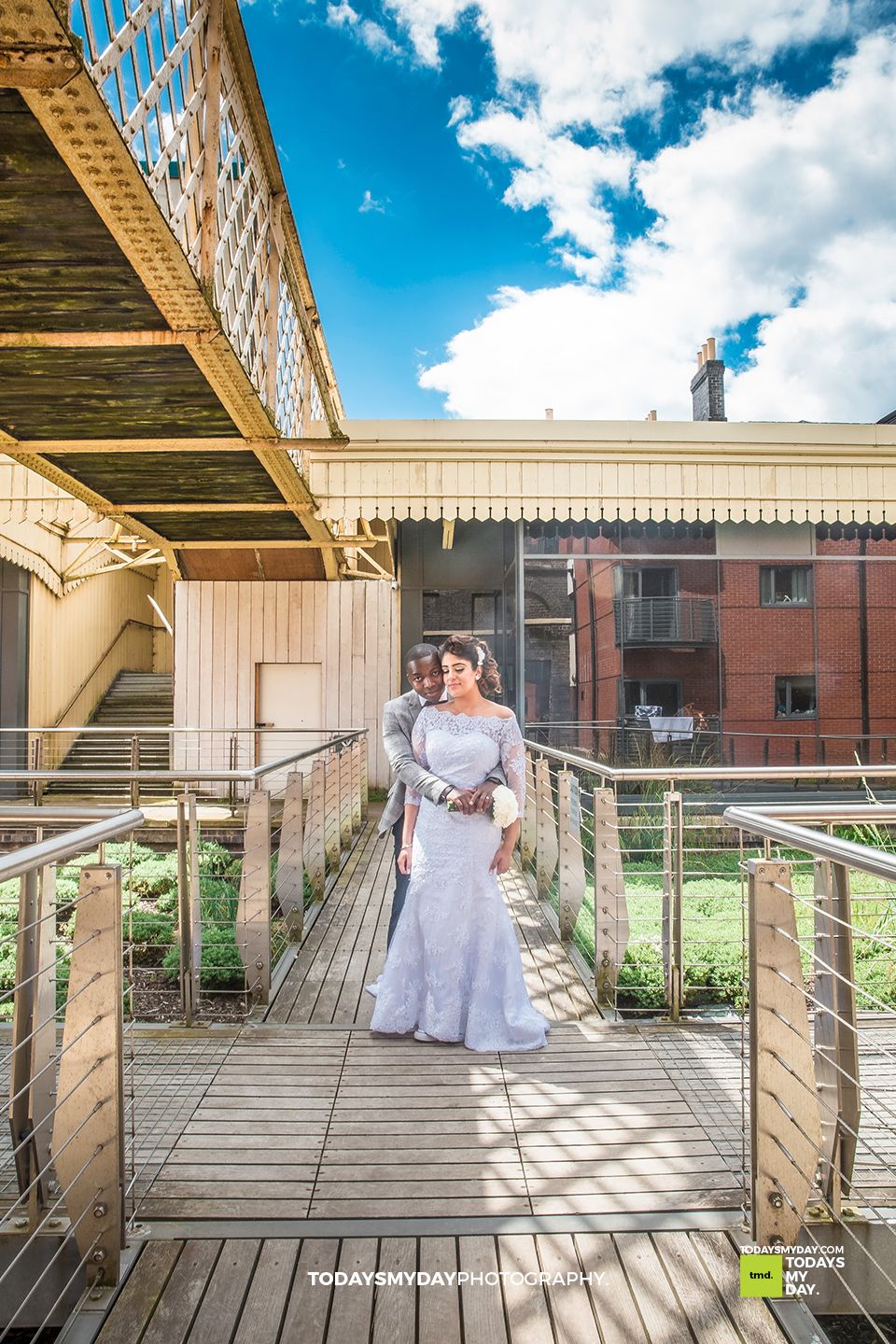 A wedding photo on a wooden bridge deck with cream colored railings against a bright blue sky.