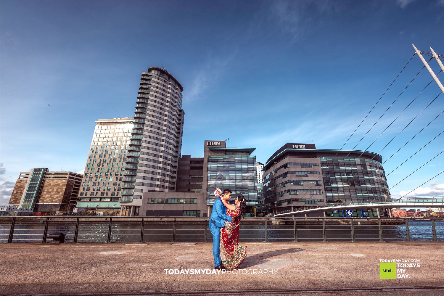 Couple in formal attire embracing in front of modern city buildings and skyline.