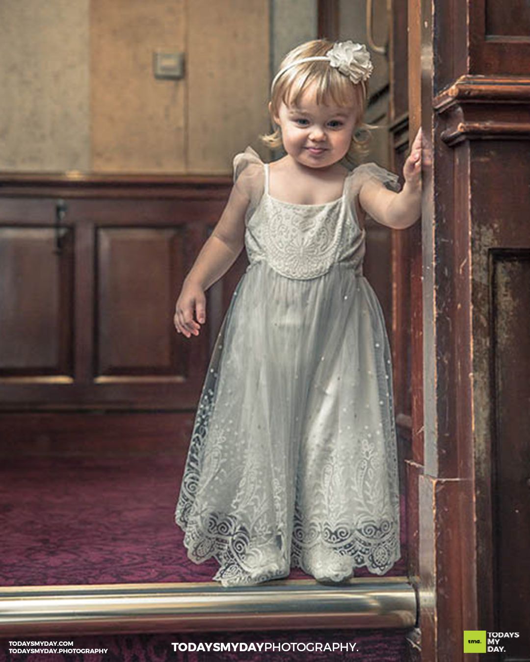 Young flower girl in white lace dress standing by wooden door in vintage setting.