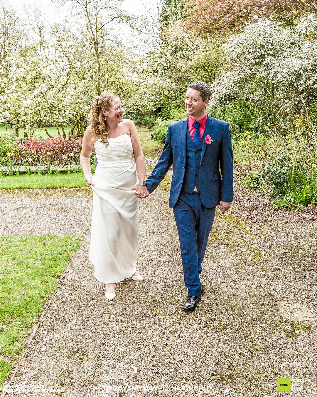 Wedding couple walking and laughing together on gravel path surrounded by spring blossoms.