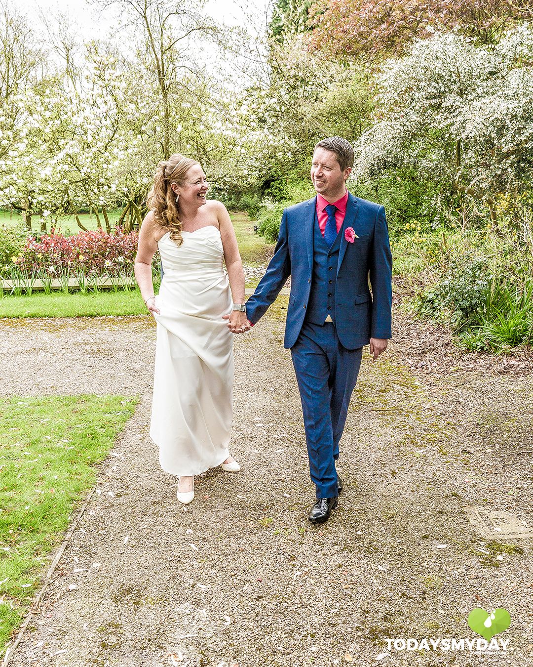 Newlyweds walking hand in hand down garden path surrounded by blooming trees.