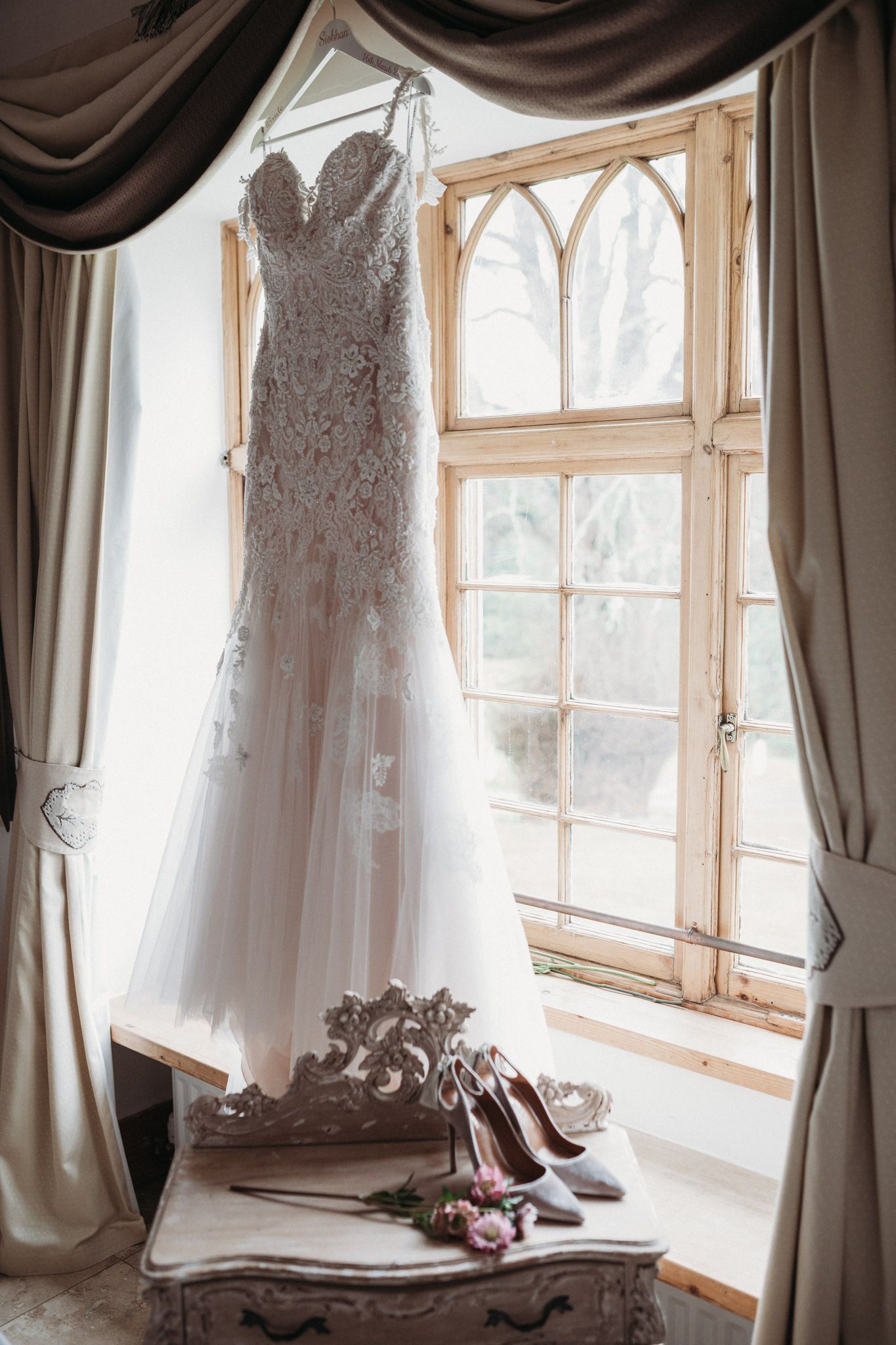 wedding dress hanging on window with bridal shoes on table