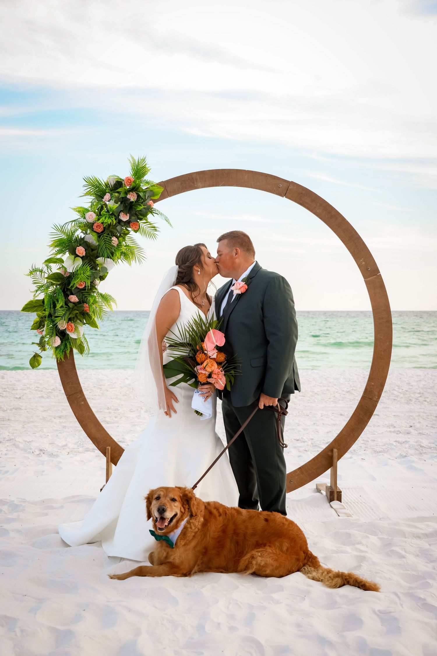 A beach wedding ceremony features a circular arch adorned with tropical flowers and a golden retriever resting on white sand.