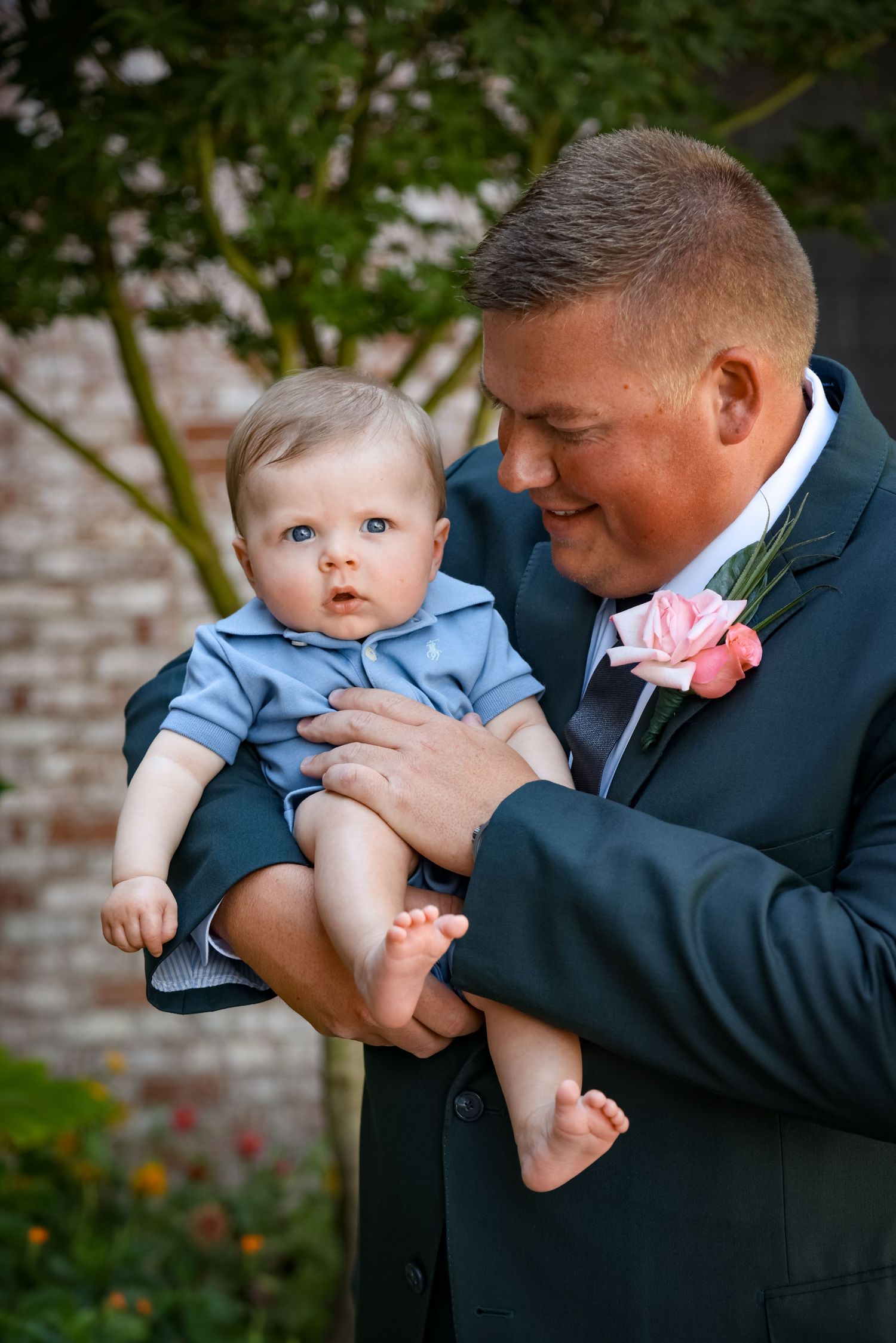 Tender moment between suited man wearing pink boutonniere and baby in blue outfit.
