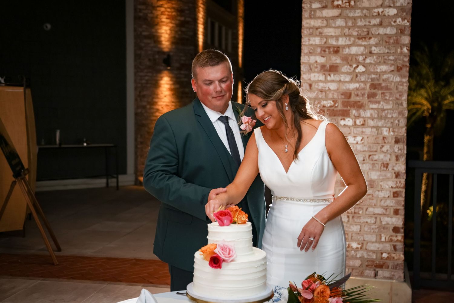 A wedding couple cuts into their elegant white and coral tiered cake in front of an exposed brick wall.
