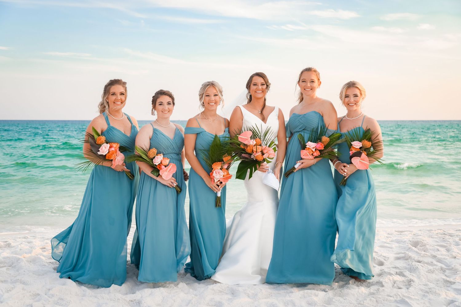 Bridesmaids in flowing turquoise dresses hold bouquets while standing on a white sandy beach.