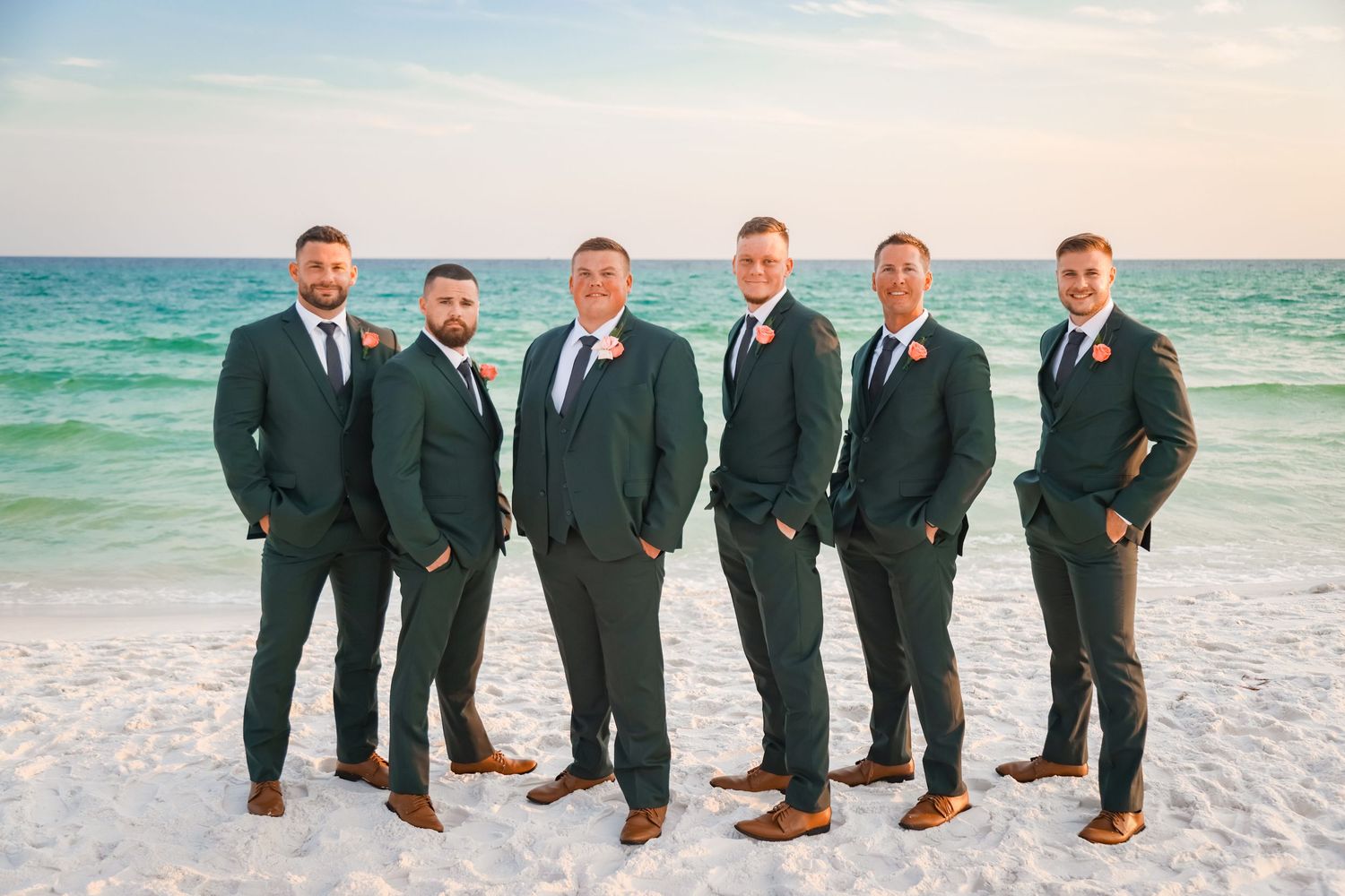 Groomsmen in charcoal suits pose together on a pristine beach with turquoise waters in the background.