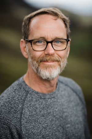 Portrait of person wearing glasses and grey sweater smiling outdoors with mountains in background.