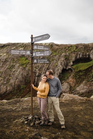 A couple poses next to a hiking trail directional sign post on a rocky mountain overlook.