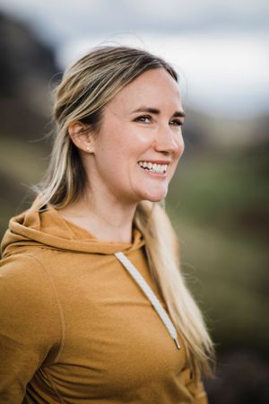 Person in mustard yellow hoodie smiles while standing on mountain trail.