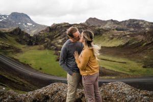 A couple shares an intimate moment on a rocky outcrop with winding roads and mountains in the background.