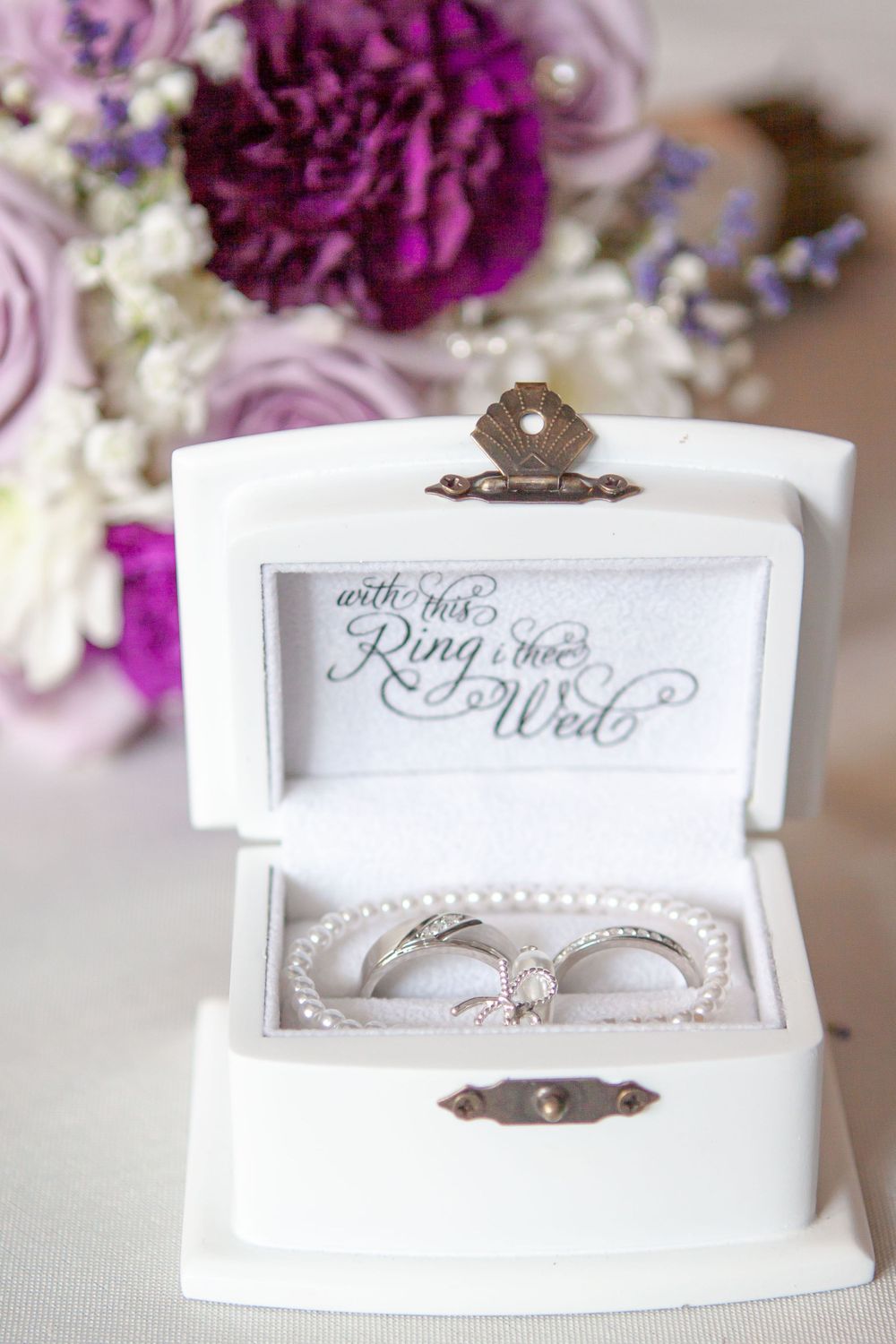 Picture of a ring box with his and hers rings inside, her bouquet in the background
