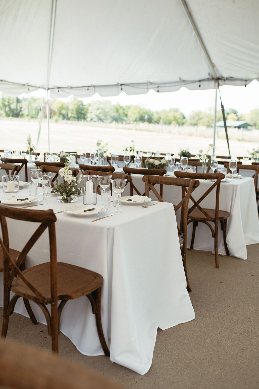 White-clothed tables with rustic wooden chairs arranged under an outdoor wedding tent.
