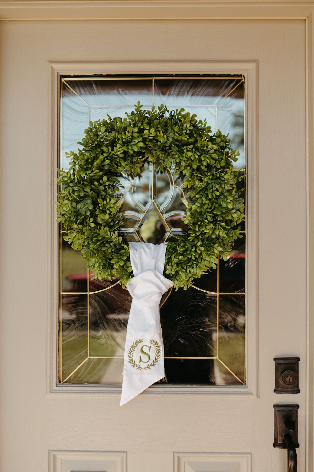Green boxwood wreath with white monogrammed ribbon hanging on beige front door.