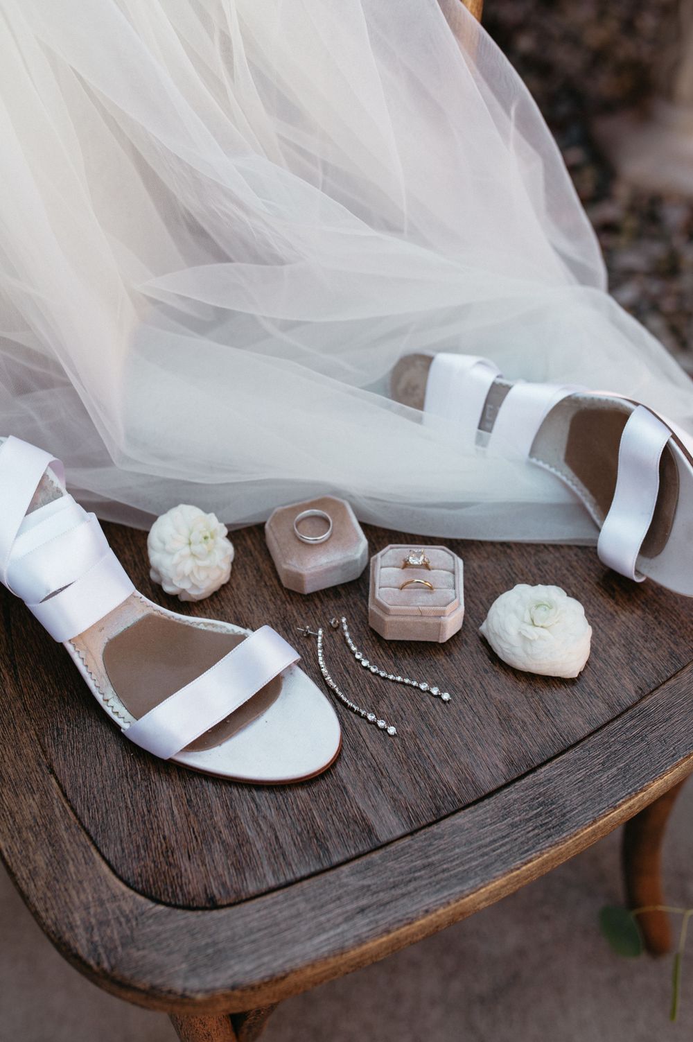 Wedding accessories including white sandals, ring boxes and jewelry displayed on dark wood surface.
