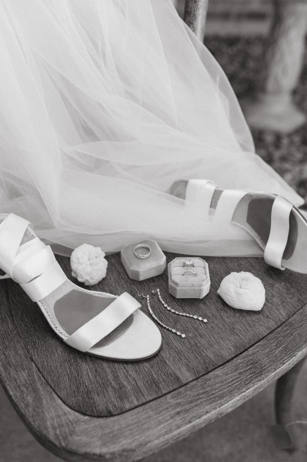 White bridal sandals with wedding accessories and ring boxes arranged on wooden chair.
