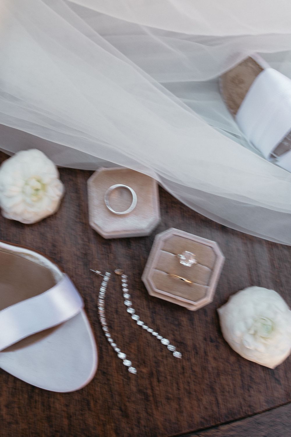 Wedding rings, jewelry and white sandals styled with tulle on dark wooden surface.