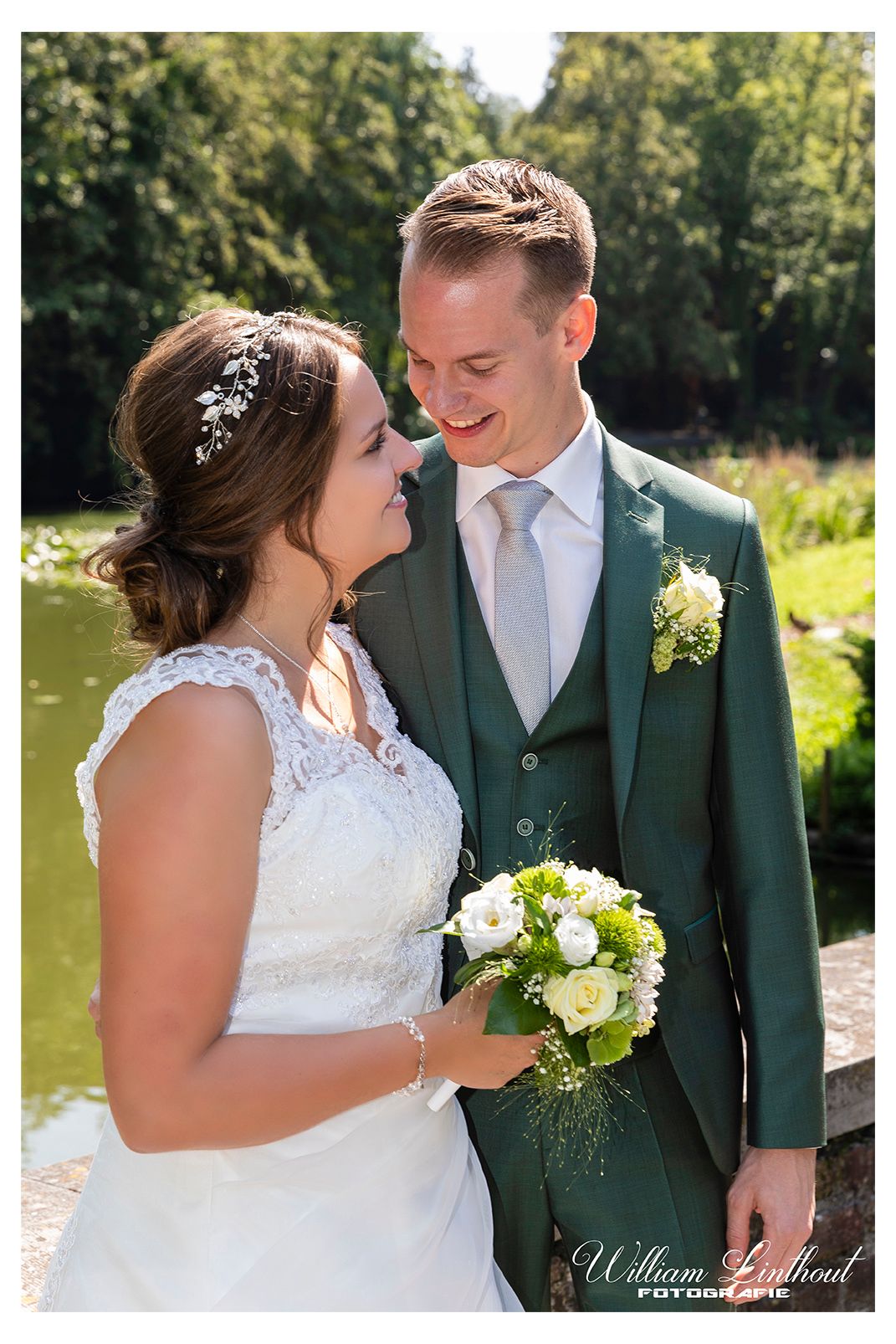 A romantic outdoor wedding photo of a couple in white dress and green suit standing by a pond.