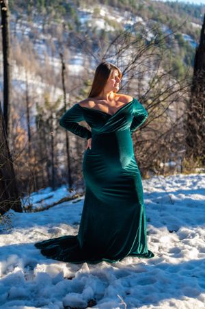 A dramatic maternity pose in an off-shoulder emerald gown surrounded by snowy winter scenery.