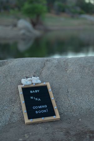 Letter board announcement with baby shoes on rocky surface near reflective water.